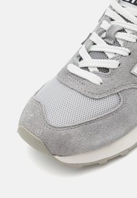 Šedá teniska s špičkou a bílou síťovinou, bílými šňůrkami a semišovým materiálem na světle šedé podešvi, zobrazená na bílém pozadí.