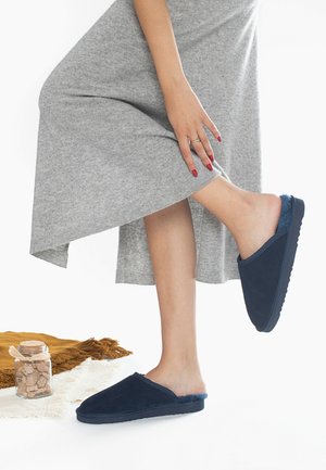 Chaussons en daim bleu marine avec une doublure en fourrure douce, design à enfiler et semelle épaisse et texturée, placés à côté d'un pot en verre sur une surface en tissu.