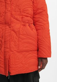 Chaqueta acolchada naranja con un patrón texturizado, cierres de cremallera y botones a presión, y un detalle de cordón ajustable en la cintura.