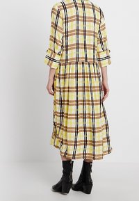 Midikleid mit einem gelben, braunen und schwarzen Karomuster, 3/4-Ärmeln und gesmocktem Taillenschnitt. Trägt dazu schwarze Knöchelstiefel.