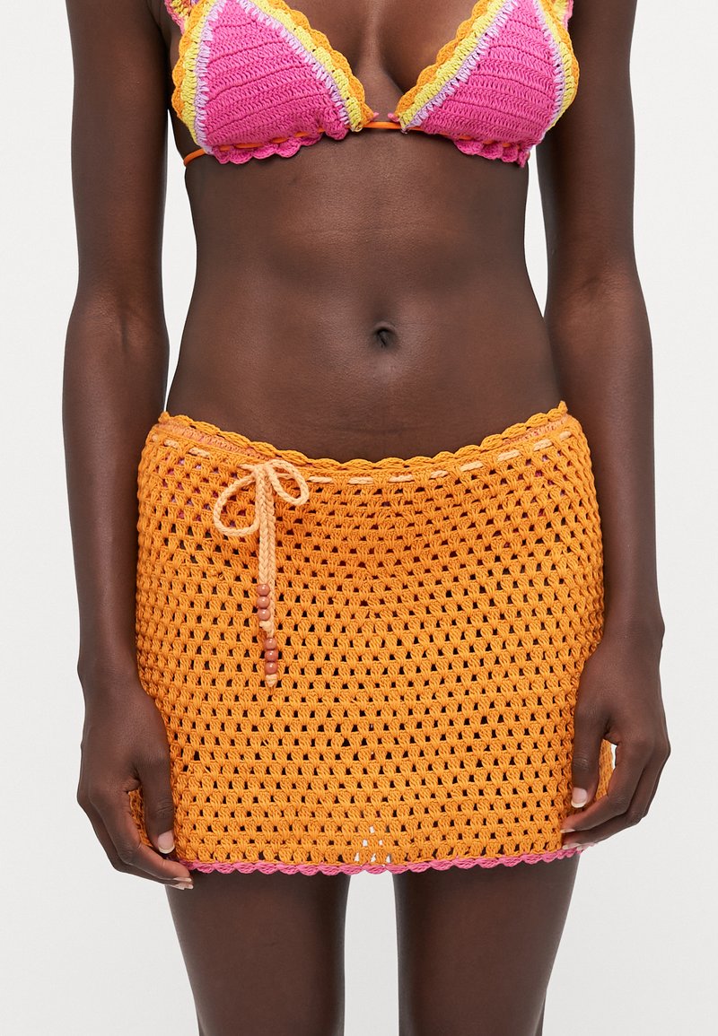 Falda de ganchillo naranja con bordes de picos rosas y detalle de lazo. Textura de tejido abierto, diseñada como para usar en la playa o como cubierta de traje de baño.