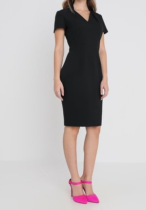 Nainen yllä mustaa muotoilevaa polvipituista mekkoa, jossa on lyhyet hihat, sekä vaaleanpunaista korkokenkää nilkkaremmillä.