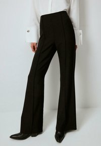 Black high-waisted trousers with a flared leg, featuring vertical seam detailing, made of a textured fabric, paired with a long-sleeve white shirt.