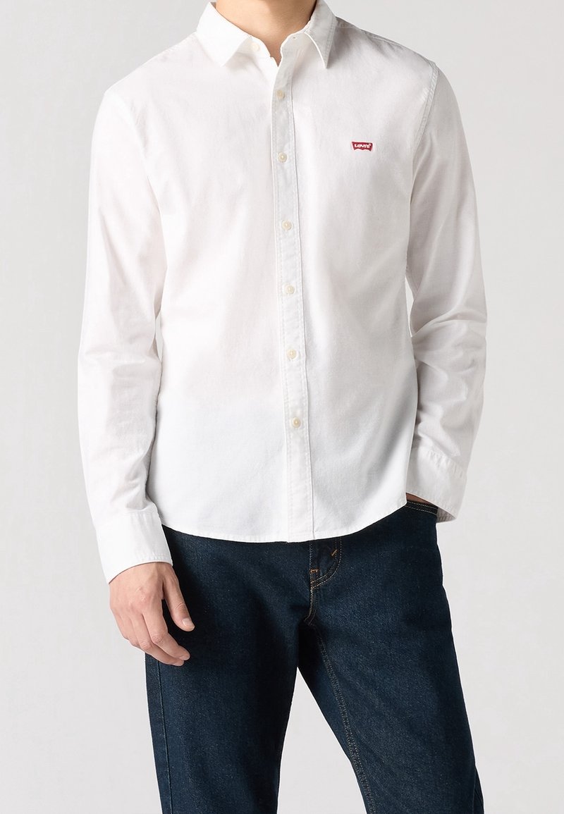 Biała koszula na guziki z długim rękawem i kołnierzykiem, z małym czerwonym logo Levi's, zestawiona z ciemnoniebieskimi jeansami.