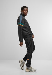 Chándal negro con acentos de color en azul, verde y naranja. Presenta una chaqueta con cremallera y cintura elástica. Zapatillas deportivas plateadas.