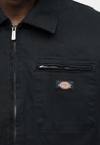 Κοντινή λήψη μαύρου μπουφάν με μεταλλικό φερμουάρ και μια μικρή τσέπη στο στήθος με φερμουάρ που εμφανίζει ένα πολύχρωμο σήμα Dickies.