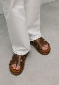 Brown slide sandals with cut-out design and contrasting stitching; worn with white pants; showing bare toes on a smooth gray surface.