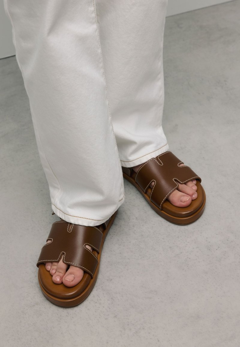 Brown slide sandals with cut-out design and contrasting stitching; worn with white pants; showing bare toes on a smooth gray surface.