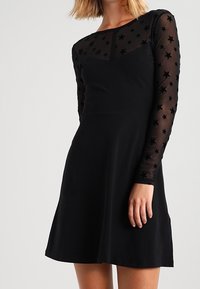 Czarna sukienka z dopasowanym gorsetem i rozkloszowanym dołem. Długie rękawy wykonane z przezroczystego materiału z wzorem gwiazd. Gładka tekstura, prosty design.