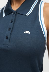 Αμάνικο πόλο μπλουζάκι σε ναυτικό μπλε με ρίγες σε λευκό και μπλε στο κολάρο και τα μανίκια, με τέσσερα κουμπιά και μικρό λευκό λογότυπο στο στήθος.