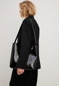 Bolso tote negro con relieve de cocodrilo y textura brillante, que presenta una sola correa para el hombro y una forma angular, sostenido por una persona con una chaqueta negra.