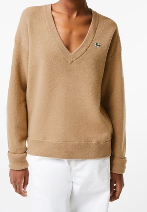 Osoba oblečená v béžovém svetru s véčkovým výstřihem a malým zeleným logem krokodýla na levé straně hrudi a bílých kalhotách, stojící s uvolněnýma rukama.