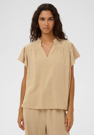 Beżowa bluzka z krótkim rękawem i dekoltem w serek, wykonana z teksturowanej tkaniny, z marszczonymi szwami na ramionach. Noszona z dopasowanymi, jasnymi spodniami.