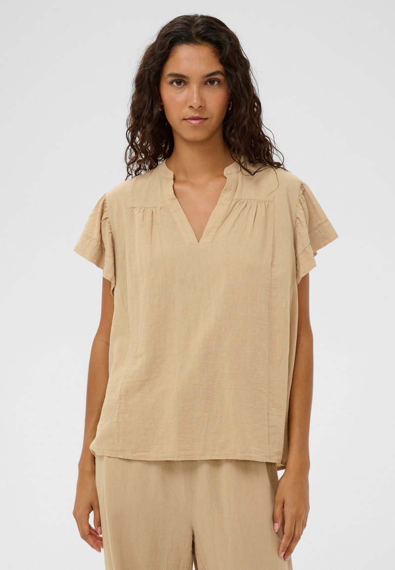 Haut beige à manches courtes avec col en V, tissu texturé et coutures froncées aux épaules. Porté avec un pantalon assorti de couleur claire.