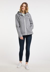 Szürke cipzáros kapucnis pulóver, zsinóros kapucnival, oldalsó zsebekkel és puha textúrával, sötét, szűk farmernadrággal és fehér sportcipővel párosítva.