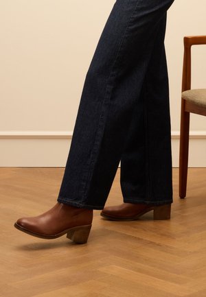 Braune Leder-Stiefeletten mit einem gestapelten Absatz, kombiniert mit dunkelblauen, weiten Jeanshosen. Im Hintergrund ist ein Holzstuhl sichtbar.