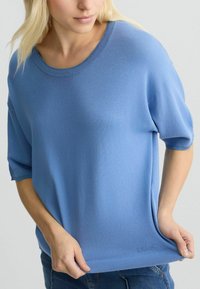 Pulover tricotat de culoare albastru deschis, cu mâneci scurte, decolteu larg și tiv ondulat. Material texturat, croi lejer și branding discret în partea de jos.