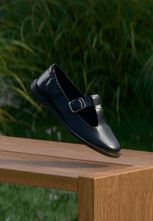 Musta nahkainen Mary Jane -kenkä hopeisella soljella leijuu puisella penkillä ulkona, taustalla vihreitä kasveja.