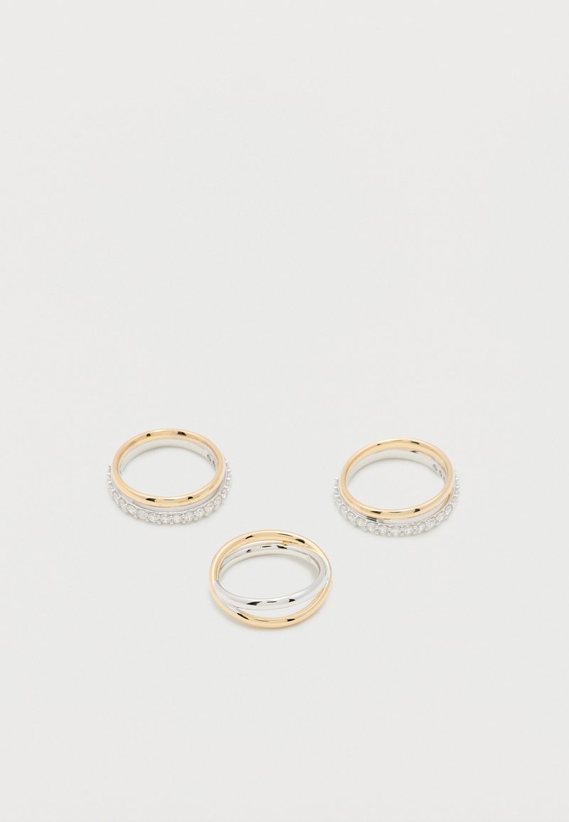 Τρία χρυσά και ασημένια δαχτυλίδια, δύο με μικρά διαμάντια γύρω από τις μπάντες, εκτίθενται σε απλή λευκή επιφάνεια.