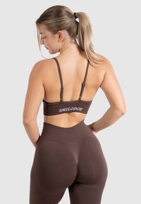 Braune nahtlose Sport-BH mit verstellbaren Trägern und Logo, kombiniert mit hochgeschnittenen braunen Leggings, die ein konturierendes Design und strukturierte Akzente aufweisen.