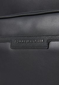 Must musta toode, millel on tekstuuriga pinnad. Käsitsemine hõlmab neljakandilist silti, millel on hõbedaste tähtedega "TOMMY HILFIGER" embosseeritud.