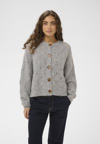 Frau mit langen, lockigen Haaren trägt eine graue, gestrickte Strickjacke mit großen Knöpfen und dunkle Hosen, steht mit einer Hand in der Tasche.