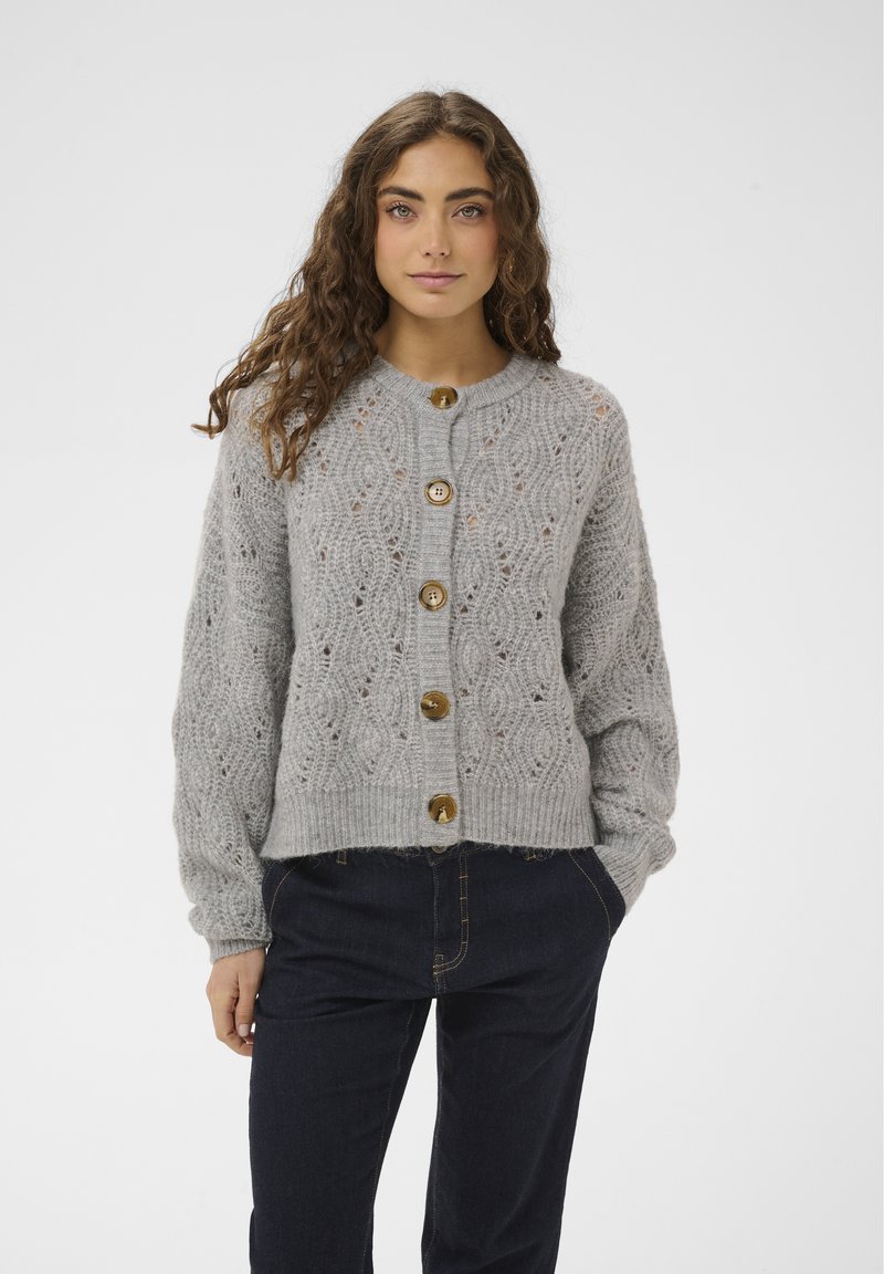 Frau mit langen, lockigen Haaren trägt eine graue, gestrickte Strickjacke mit großen Knöpfen und dunkle Hosen, steht mit einer Hand in der Tasche.