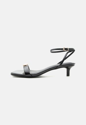 Μαύρο δερμάτινο σανδάλι με ανοιχτά δάχτυλα, χαμηλό τακούνι kitten, λεπτό λουράκι στον αστράγαλο με χρυσή αγκράφα και διακοσμητική χρυσή λεπτομέρεια στο μπροστινό λουράκι.