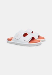 Sandale albe cu talpă plată, dotate cu două curele reglabile din Velcro. Pad-ul pentru picior este portocaliu, iar talpa este albă, având un model texturat pentru aderență.