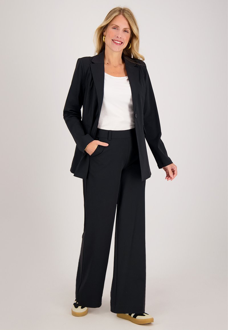 Zwarte blazer met één knoop, gecombineerd met zwarte pantalon met wijde pijpen en een wit topje. Heeft een zachte stof en een relaxte pasvorm.