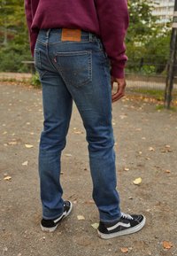 Jean en denim bleu avec une coupe droite, doté d'un patch en cuir marron à la ceinture arrière et de poches arrière avec des coutures contrastantes.