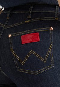 Πίσω τσέπη τζιν από σκουρομπλε τζιν με κίτρινη ραφή, χαλκομανίες από χαλκό και κόκκινη ετικέτα της μάρκας Wrangler.