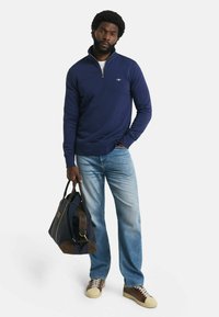 Uomo che indossa un maglione a zip blu navy, jeans azzurri chiari, sneakers marroni, mentre tiene una borsa a tracolla blu navy e marrone contro uno sfondo bianco.