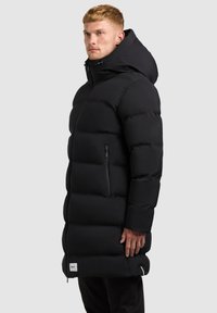 Lang sort dunjakke med hætte, der har en lynlåslomme på brystet, tekstureret overflade og en justerbar snøre til hætten.