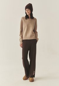 Maglione beige con design di orso in rilievo, polsini a costine, abbinato a pantaloni marroni a vestibilità ampia e scarpe slip-on in pelle scamosciata color sabbia. Berretto marrone.