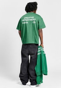 Camiseta verde de manga corta con "DEPARTAMENTO CREATIVO 2018 CD" impreso en blanco en la parte posterior. Jeans negros holgados, sosteniendo una sudadera con capucha verde a juego.