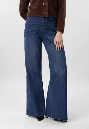 Γυναίκα που φοράει μπλε τζιν παντελόνι με ψηλή μέση και φαρδιά μπατζάκια με ξεφτισμένα τελειώματα, συνδυασμένο με καφέ κουμπωτό πουλόβερ από χνουδωτό ύφασμα και μαύρα μυτερά παπούτσια.