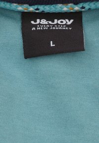 Licht turquoise stoffen label met het "J&JOY" logo, verzorgingsinstructies en maat "L." De stof heeft een ribbelstructuur met contrasterende marineblauwe accenten.
