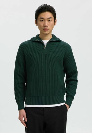 Homme portant un pull en maille côtelée vert foncé avec un col à demi-zip, un sous-maillot blanc et un pantalon noir, debout devant un fond clair.