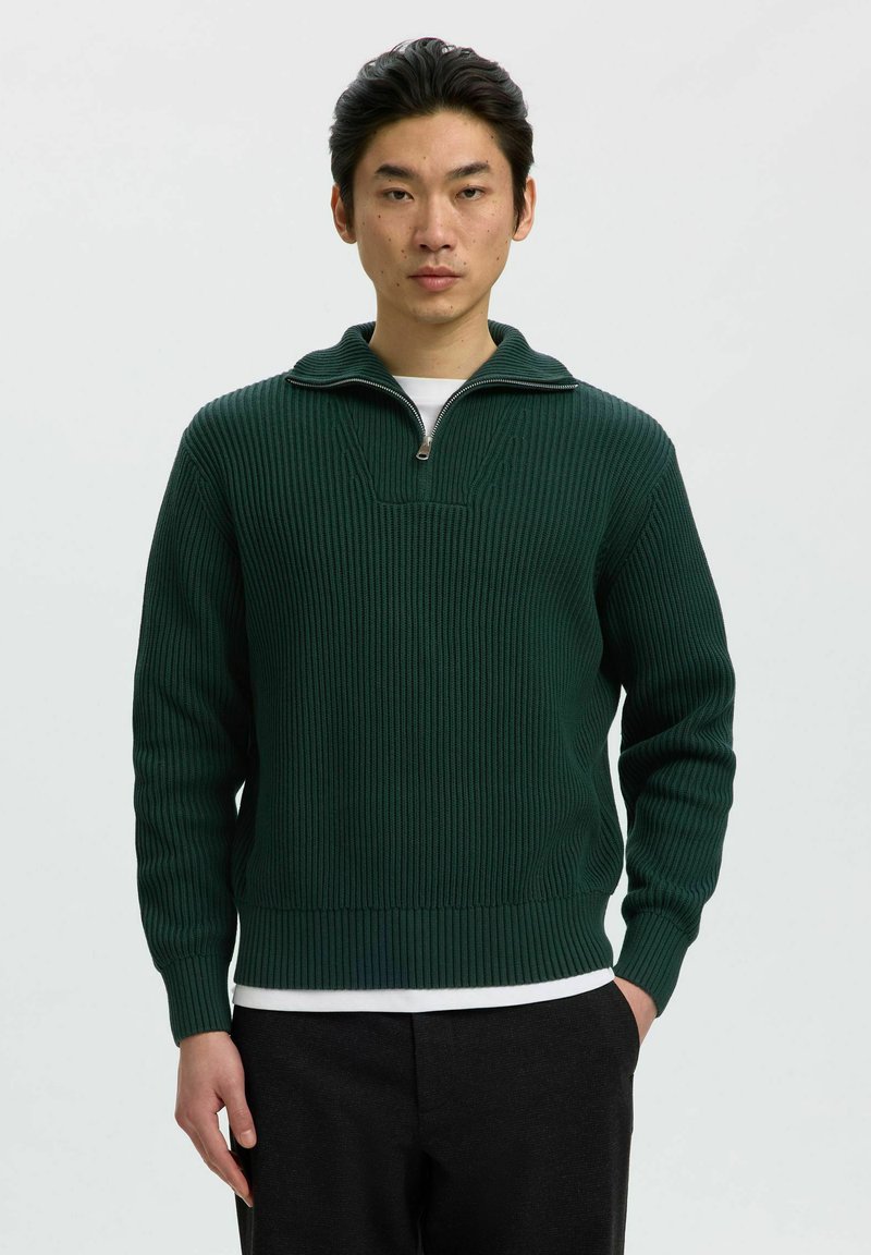 Homme portant un pull en maille côtelée vert foncé avec un col à demi-zip, un sous-maillot blanc et un pantalon noir, debout devant un fond clair.