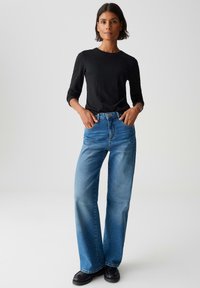 Frau mit kurzem, dunklem Haar, die ein schwarzes Langarmshirt, blaue hochtaillierte, weite Jeans und schwarze Schuhe trägt und mit den Händen in den Taschen steht.