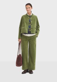 Grüne Cordjacke mit Knopfverschluss, kombiniert mit grünen Cordweite-Hosen. Das Modell trägt eine burgunderfarbene Wildledertasche.