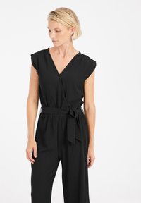 Zwarte jumpsuit met korte cap mouwen, V-halslijn en een strikriem in de taille. Gemaakt van een textuurstof, met losse, wijde pijpen.