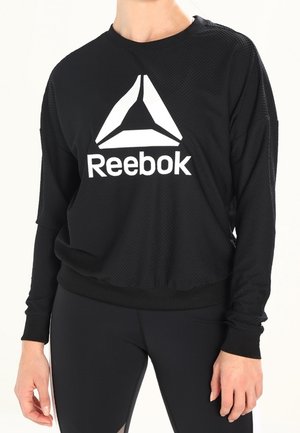 Person trägt ein schwarzes langärmeliges Reebok-Shirt mit großem weißem Logo und schwarzen Leggings vor einfachem Hintergrund.