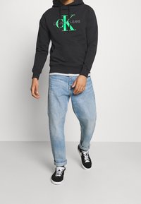 Felpa nera con logo "CK" verde, jeans in denim blu chiaro e sneakers nere con suola bianca. Stile casual, vestibilità rilassata.