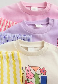 Tres sudaderas en rosa, morado y crema, con patrones y detalles juguetones, combinadas con pantalones a rayas amarillas, todo de algodón.