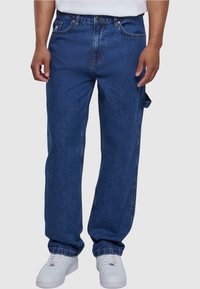 Jeans de denim azul con un corte relajado, cinco bolsillos y una pequeña correa lateral. Combinados con zapatillas blancas, que presentan un diseño clásico.