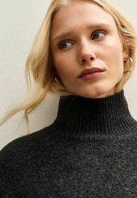 Blonde vrouw met blauwe ogen draagt gouden creolen en een donkergrijze geribbelde coltrui, kijkt iets opzij.