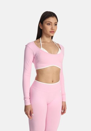 Mladá žena nosiaca svetloružový crop top s dlhým rukávom a zodpovedajúce legíny s vysokým pásom, pózujúca na jednoliatom bielom pozadí.