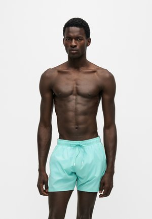Homme musclé à la peau foncée debout, portant un short de bain turquoise clair sur un fond clair uni avec une expression sérieuse.